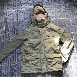 Patagonia Grön jacka - Säljer en grön jacka från Patagonia med avtagbar huva och mjukt foder. Jackan har dragkedja och knappar framtill samt flera praktiska fickor. Perfekt för kyligare dagar.