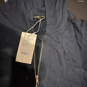 Mörkblå väst med dragkedja från LMTD - Snygg mörkblå väst från LMTD med dragkedja och huva. Perfekt för lager-på-lager-stil. Västen har en enkel design med en liten logga på bröstet.