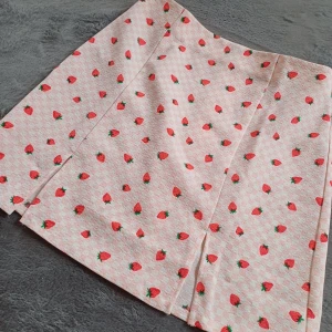 Rutig kjol med jordgubbsmönster från Monki - Söt kortkjol från Monki med ett lekfullt jordgubbsmönster på en rosa och vit rutig bakgrund. Kjolen har en slits framtill som ger en extra touch. Perfekt för en somrig look! 🍓