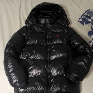 Svart dunjacka från Polo Ralph Lauren - Säljer en snygg svart dunjacka från Polo Ralph Lauren. Jackan har en quiltad design med dragkedja och huva. Perfekt för kyliga dagar med sin stilrena look och varma fyllning.