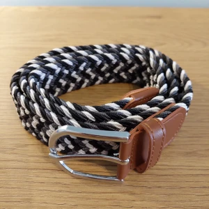 Flätat bälte med läderdetaljer - Snyggt flätat bälte i svart, vitt och grått med bruna läderdetaljer. Bältet har ett silverfärgat spänne och en unik design som ger en stilren look. Perfekt för att ge din outfit det lilla extra.