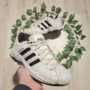 Vita sneakers från Adidas - Snygga vita sneakers från Adidas med svarta detaljer och klassiska tre ränder på sidan. Skorna har snörning och en bekväm passform, perfekt för vardagsbruk. Den stilrena designen gör dem lätta att matcha med olika outfits.
