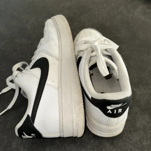 Nike Air Force i vitt och svart - Säljer ett par klassiska Nike Air Force sneakers i vitt med svarta detaljer. Skorna har snörning och den ikoniska Nike-loggan på sidan. Perfekta för en stilren look.