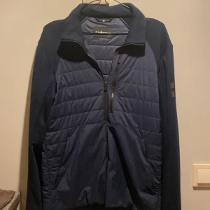 Mörkblå cardigan från Sail Racing - Säljer en mörkblå jacka från Sol's med quiltad design och dragkedja framtill. Jackan har långa ärmar och en stilren look med ett diskret märke på ärmen. Perfekt för kyligare dagar.