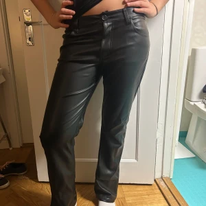 Svarta skinnbyxor - Snygga svarta skinnbyxor med låg/midwaist och rak passform. Perfekta för en stilren look. Byxorna har en klassisk design med knappar och dragkedja framtill. Inga fickor baktill