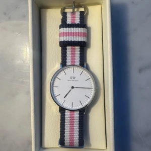 Klocka från Daniel Wellington - Säljer en stilren klocka från Daniel Wellington med en vit urtavla och ett randigt armband i blått, rosa och vitt. Klockan har en minimalistisk design med silverfärgad boett. Perfekt för den som gillar en klassisk och tidlös stil. Helt ny, aldrig använd🥰