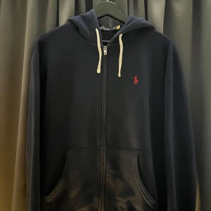 Mörkblå hoodie från Ralph Lauren - OBS LJUSET BLEV SKUMT DEN ÄR MÖRKARE NÄR MAN KOLLAR MED ÖGAT! Säljer en snygg mörkblå hoodie från Ralph Lauren med dragkedja och röd logga på bröstet. Den har en justerbar huva med vita snören och praktiska fickor framtill. Perfekt för en avslappnad stil.