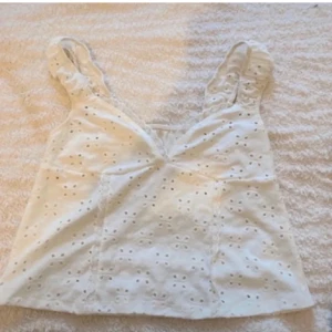 Vit ärmlös topp med spetsdetaljer - Säljer en söt vit ärmlös topp med spetsdetaljer och broderade hålmönster. Toppen har en v-ringad design och volangdetaljer vid axlarna. Perfekt för varma sommardagar! 🌼