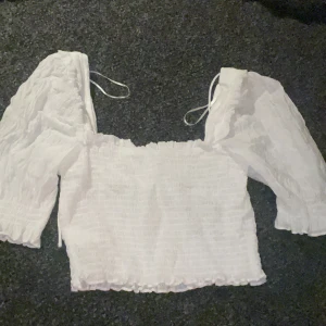 Vit blus - Säljer en söt vit blus från H&M med puffiga trekvartsärmar och smockdetaljer. Blusen har knytband vid axlarna för en justerbar passform. Perfekt för en somrig look💞