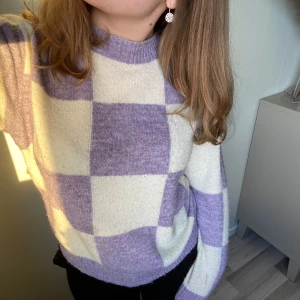 Lila och vit rutig stickad tröja - Säljer en mysig stickad tröja i lila och vitt med ett rutigt mönster. Tröjan har långa ärmar och en rund halsringning. Perfekt för kyliga dagar!