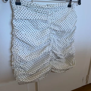Vit prickig kjol - Säljer en söt vit kjol med svarta prickar. Kjolen har en rynkad design som ger en fin struktur och form. Perfekt för en somrig look!