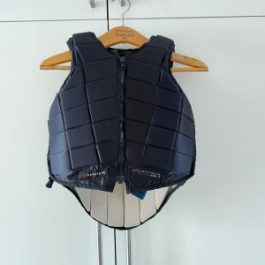 Svart säkerhetsväst från Racesafe - Säljer en svart säkerhetsväst från Racesafe. Västen har en quiltad design med dragkedja framtill och justerbara sidor. Den är i bra skick med lite "knotter" från tvätt på insidan.  Den är i storlek children L och ryggen är i x-long.