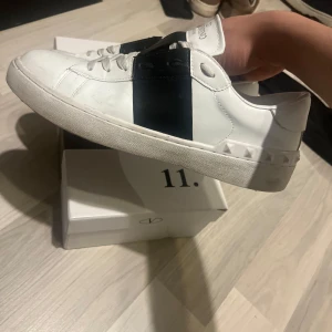 Valentino sneakers med svart detalj - Snygga vita sneakers från Valentino med en bred svart rem över ovansidan. Skorna har en klassisk design med snörning och små nitar runt hälen för en extra touch. Perfekta för en stilren look. Pris går såklart att diskutera 