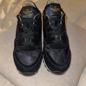 Svarta sneakers från Valentino Garavani - Snygga svarta sneakers från Valentino Garavani med coola nitar runt hälen och sulan. Skorna har en stilren design med snörning och en diskret camouflage-mönster. Perfekta för att ge din outfit en lyxig touch. Priset kan diskuteras 