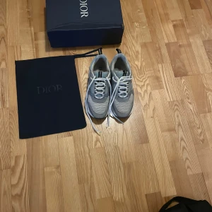 Grå och vita sneakers från Dior - Säljer ett par stilrena grå och vita sneakers från Dior. Skorna har en modern design med snörning och en bekväm sula. Perfekta för dig som vill ha både stil och komfort. Levereras med originalkartong och påse.