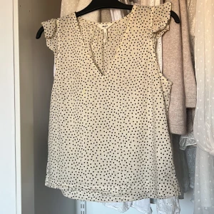 Beige prickig blus - Säljer en söt beige blus med svarta prickar. Blusen har en v-ringad design och korta volangärmar som ger en romantisk touch. Perfekt för både vardag och speciella tillfällen.