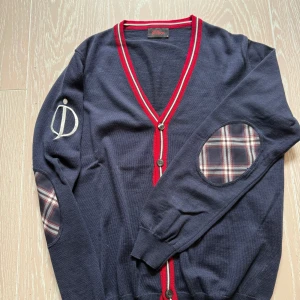 Mörkblå kofta från Oscar Jacobson - Snygg mörkblå kofta från Oscar Jacobson med röda och vita detaljer. Den har knappar framtill och stiliga rutiga armbågslappar. Perfekt för en stilren look!