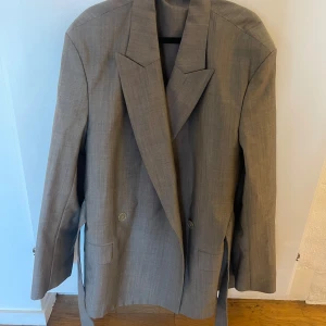 Grå kostym från Acne Studios - Snygg kostym från Acne Studios i grått. Jackan har en dubbelknäppning och en bälte i midjan. Byxorna har en elastisk midja. Kostymen är knappt använd, så den är i riktigt fint skick. Storlek 34 i kavajen och byxorna. Kostymen är oversize. Rökfritt hem. Skickar paket som spårbart.