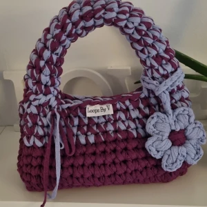 Virkad väska från Loops By V - Säljer en unik virkad handväska från Loops By V i en härlig kombination av ljuslila och cyren. Väskan är virkad I dubbelt bandgarn av återvunnen bomull och har en avtagbar blomma. Perfekt för att ge din outfit en personlig touch! En robust modell som sticker ut.