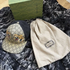 Gucci tiger keps  - Snygg keps från Gucci med ikoniskt GG-mönster och broderade tigrar framtill. Kepsen har en svart mesh-bakdel och justerbart band med klassiska Gucci-färger. Perfekt för att ge din outfit en lyxig touch.