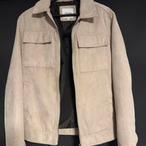 Beige mockajacka från Jack & Jones - Snygg beige mockajacka från Jack & Jones i storlek S. Jackan har en klassisk krage och två bröstfickor med lock. Perfekt inför våren.