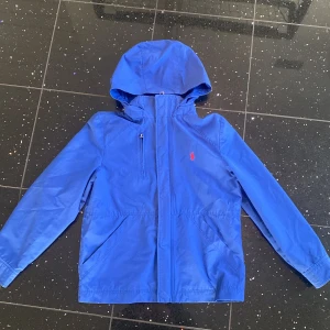 Blå vindjacka från Polo Ralph Lauren - Säljer en snygg blå vindjacka från Polo Ralph Lauren. Jackan har en huva och dragkedja framtill. Perfekt för blåsiga dagar och har en stilren design med det klassiska Polo-logotypen på bröstet.