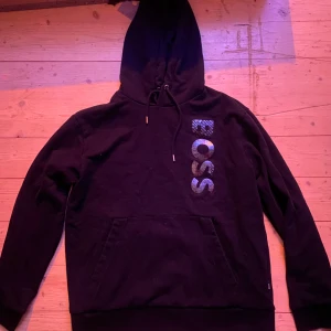 Svart hoodie från Boss - Snygg svart hoodie från Boss med stor logga på framsidan. Den har en klassisk design med huva och dragsko samt en praktisk magficka. Perfekt för en avslappnad stil.