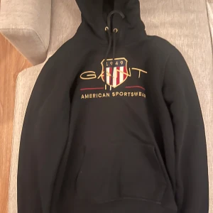 Svart hoodie från GANT - Säljer en svart hoodie från GANT med ett stort broderat emblem på bröstet. Hoodien har en justerbar huva med snören och en stor ficka framtill. Perfekt för en avslappnad stil.