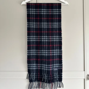 Halsduk från Burberry - Snygg och klassisk rutig halsduk från Burberry i svart, grått och rött. Tillverkad i mjuk kashmir med fransar i ändarna. Perfekt för att hålla dig varm och stilren under kyliga dagar.