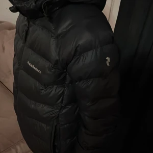 Svart dunjacka från Peak Performance - Säljer en svart dunjacka från Peak Performance. Jackan har en pufferdesign med en justerbar huva och dragkedja framtill. jackan används för sommar och vår. En liten skada på jackan vid luvan men inget farligt.😊