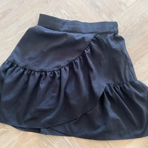 Svart omlottkjol från Lindex - Snygg svart omlottkjol från Lindex i återvunnen polyester. Kjolen har volanger som ger en fin detalj och en feminin touch. Perfekt för att skapa en stilren look.