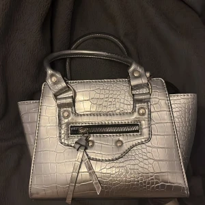 Silvrig handväska med krokodilmönster - Snygg silvrig handväska med krokodilmönster och dragkedja framtill. Väskan har två handtag och en dekorativ rem med knytning. Perfekt för att ge din outfit en glammig touch. Medföljer axelremsband