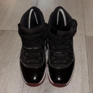 Jordan 11 bred - Snygga Nike Air Jordans i svart och rött med hög design och snörning. Skorna har en glansig finish och en vit mellansula. Perfekta för den som vill ha en stilren och sportig look.