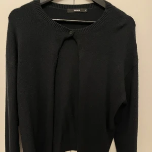 Svart kofta från Bik Bok - Snygg svart kofta från Bik Bok med rund halsringning och knappdetalj framtill. Perfekt för lager-på-lager och att hålla sig varm under kyligare dagar.