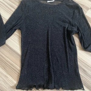Svart glittrig tröja - Snygg svart glittrig tröja genom skinlig med långa ärmar. Perfekt för en kväll ute eller en festlig tillställning. Tröjan har en enkel design med en rund halsringning och en lätt vågig kant vid ärmslut och nederkant.