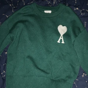 Grön stickad tröja från Ami - Säljer en snygg grön stickad tröja från Ami med ett vitt hjärta och bokstaven A på bröstet. Tröjan har en rund halsringning och ribbade kanter vid ärmslut och nederkant. Perfekt för en stilren look!