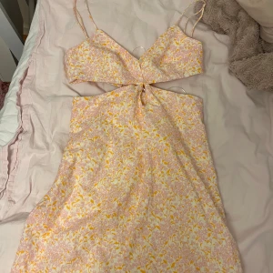 Blommig klänning med cut-out detaljer - Söt blommig klänning i ljusa toner med smala axelband och en trendig cut-out detalj framtill. Perfekt för sommarens varma dagar. Klänningen har en v-ringad topp och en lätt utsvängd kjol.