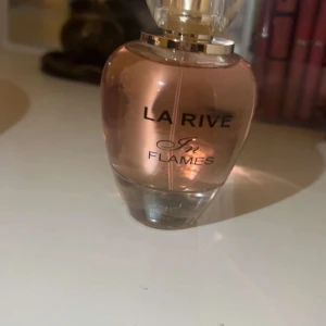 La Rive In Flames Parfym - Elegant parfymflaska från La Rive med namnet 'In Flames'. Flaskan har en rundad form och är i en mjuk rosa nyans med en transparent bas. Korken är genomskinlig och ger en lyxig känsla. Perfekt för den som söker en sofistikerad doftupplevelse.