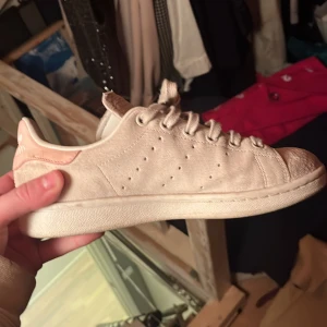 Adidas Stan Smith i beige mocka - Snygga Adidas Stan Smith sneakers i beige mocka med klassisk design. Skorna har en ljusrosa detalj på hälen och perforerade ränder på sidorna. Perfekta för en stilren look.
