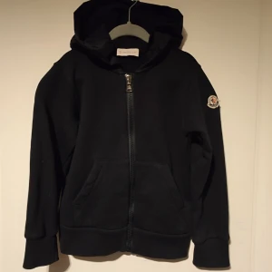 Svart huvtröja från Moncler - Säljer en svart huvtröja från Moncler med dragkedja och fickor framtill. Tröjan har en stor logga på huvan och ribbade muddar vid ärmslut och nederkant. Perfekt för en stilren look. 