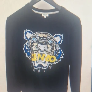 Svart tröja med tigerbroderi från Kenzo - Säljer en svart tröja från Kenzo med ett stort, färgglatt tigerbroderi på framsidan. Tröjan har långa ärmar och en rund halsringning. Perfekt för att ge din outfit en cool touch 🐯.160-166 cm skulle passa dej