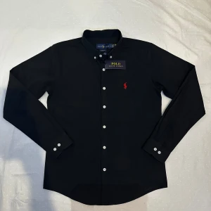 Svart skjorta från Ralph Lauren - Snygg svart skjorta från Ralph Lauren med klassisk krage och knappar framtill. Den har en broderad logotyp i rött på bröstet och är tillverkad i 100% bomull. Perfekt för en stilren look.