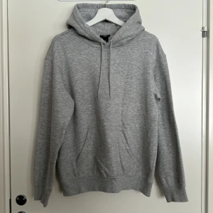 Grå hoodie från H&M - Säljer en stilren grå hoodie från H&M i relaxed fit. Den har en klassisk design med justerbar huva och en stor magficka framtill. Perfekt för en avslappnad look. Inga defekter.