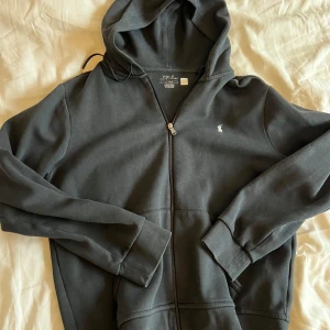 Svart hoodie från Ralph Lauren - Säljer en stilren svart hoodie från Ralph Lauren med dragkedja och huva. Tröjan har långa ärmar och en liten logotyp på bröstet. Perfekt för en avslappnad look. Använd 3 gånger.