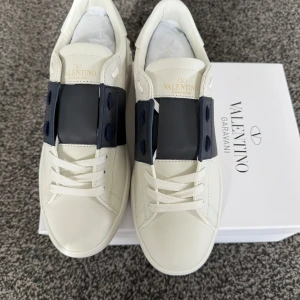 Valentino Garavani sneakers i marinblå - Snygga Valentino Garavani sneakers i vitt med en bred blå rem över ovansidan. Skorna har en stilren design med snörning och är tillverkade i skinn. Perfekta för en trendig look.