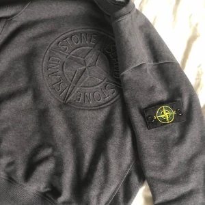 Grå sweatshirt från Stone Island - Säljer en stilren grå sweatshirt från Stone Island med deras ikoniska logga broderad på bröstet och en patch på ärmen. Tröjan har rund hals och ribbade muddar vid ärmslut och nederkant. Perfekt för en casual look. Tröjan tvättas och strycks innan jag skickar den. Skickas inom 2 dagar efter köp🔥 Pris ej hugget i sten🪨🪓. Skriv gärna om du har frågor mvh, Linus😃