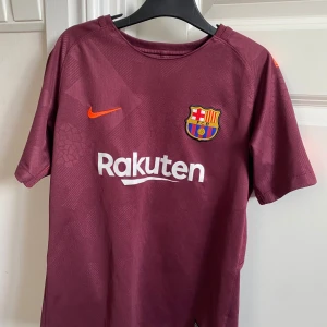 Vinröd FC Barcelona fotbollströja från Nike - Säljer en vinröd FC Barcelona fotbollströja från Nike med korta ärmar. Tröjan har klubbens emblem och sponsorlogga på framsidan samt spelarnamnet 'Coutinho' och nummer 14 på baksidan. Tillverkad i Dri-FIT-material för optimal komfort.