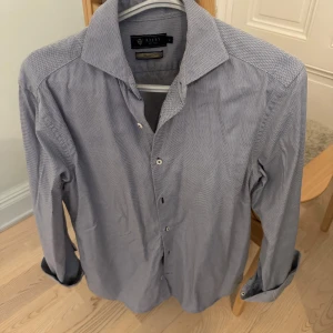 Blå långärmad skjorta från Riley - Snygg blå långärmad skjorta från Riley med ett diskret mönster. Skjortan har knappar framtill och en klassisk krage. Perfekt för en stilren look.