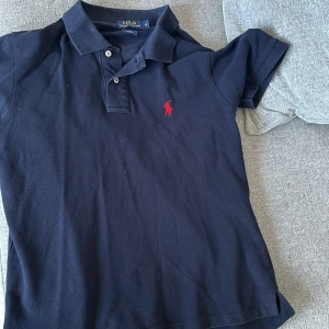 Blå pikétröja från Ralph Lauren - Säljer en klassisk svart pikétröja från Ralph Lauren med den ikoniska röda loggan på bröstet. Tröjan har en krage med knappar och korta ärmar. Perfekt för en stilren look.