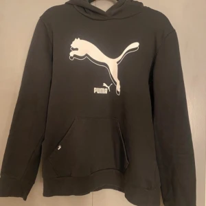 Svart hoodie från Puma - Säljer en svart hoodie från Puma med det klassiska logotyptrycket i vitt på framsidan. Tröjan har en stor ficka framtill och en bekväm huva. Perfekt för en avslappnad stil.